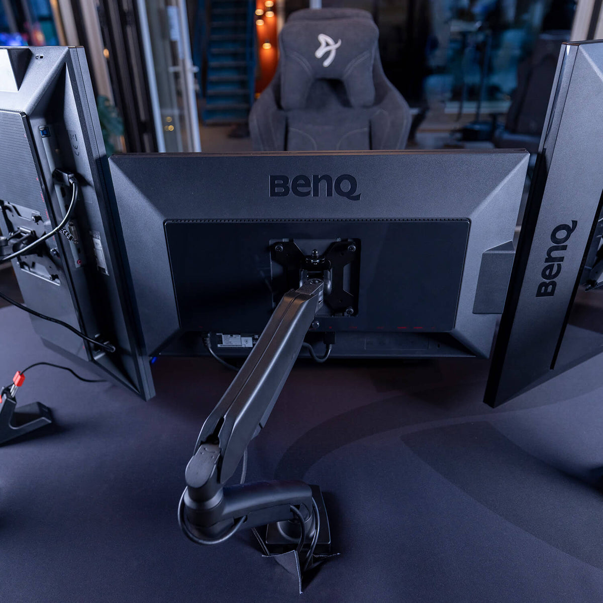A gaming setup featuring two BenQ monitors mounted on adjustable arms, viewed from behind, with a gaming chair and part of a modern room visible in the background.