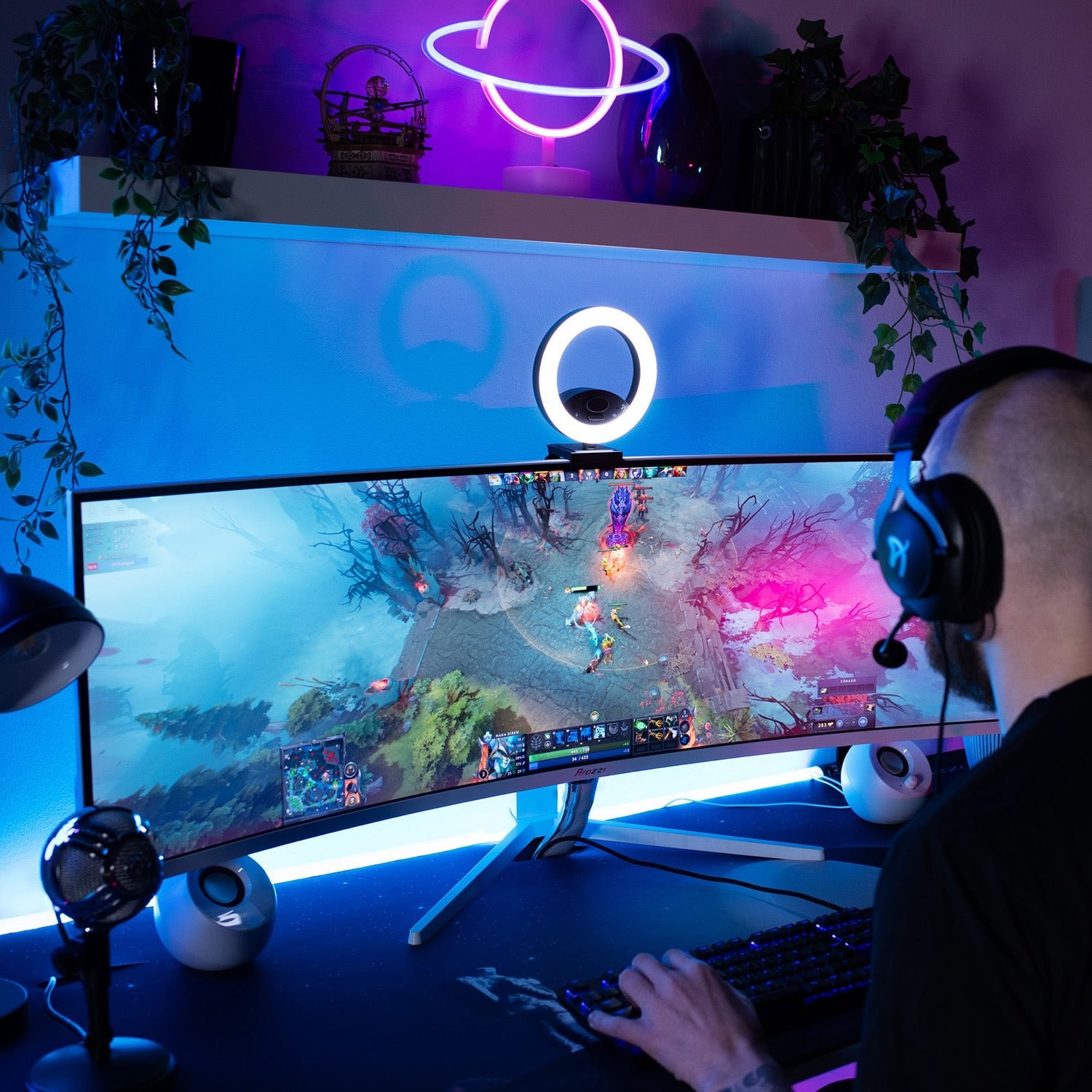 Wearing headphones, a person plays a vibrant video game on the Arozzi Nova 49″ Curved VA 165Hz Ultrawide Gaming Monitor, which features adaptive sync technology and is set up in an immersive environment with blue and purple LED lights, lush plants, and a glowing neon planet sign on the wall.