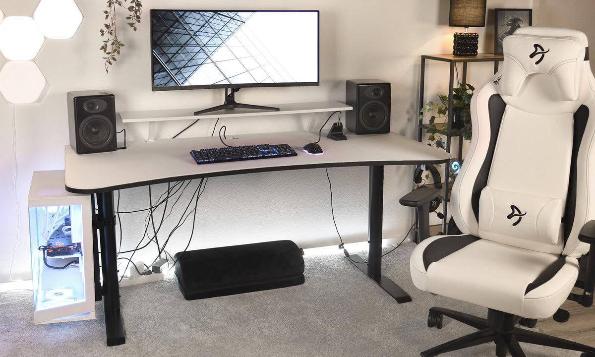 A modern gaming setup with a large monitor, keyboard, mouse, speakers, and a white gaming chair. A PC with visible components sits on the floor beside the desk. Cables hang under the desk.
