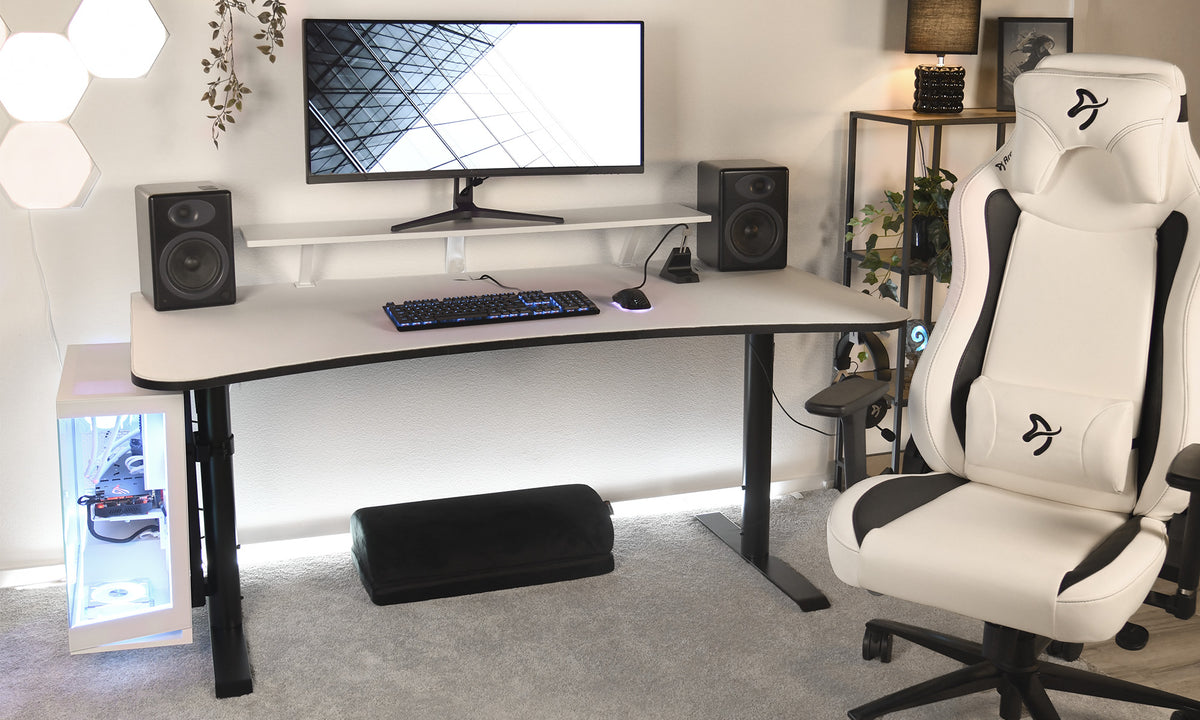 A modern gaming setup with a white desk, black speakers, a large monitor, RGB keyboard, mouse, white gaming chair, footrest, and a glass-sided PC tower with blue lighting. Minimalist decor and neutral tones.