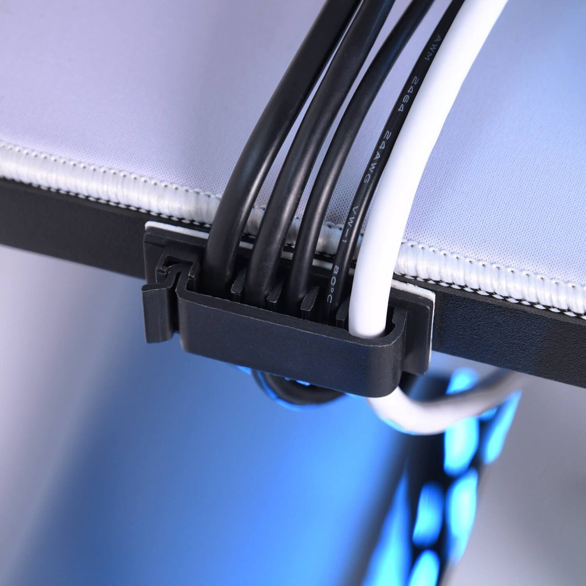 Close-up of a cable management clip attached to the edge of a desk, organizing several black and white cables. Blue light glows underneath the desk.