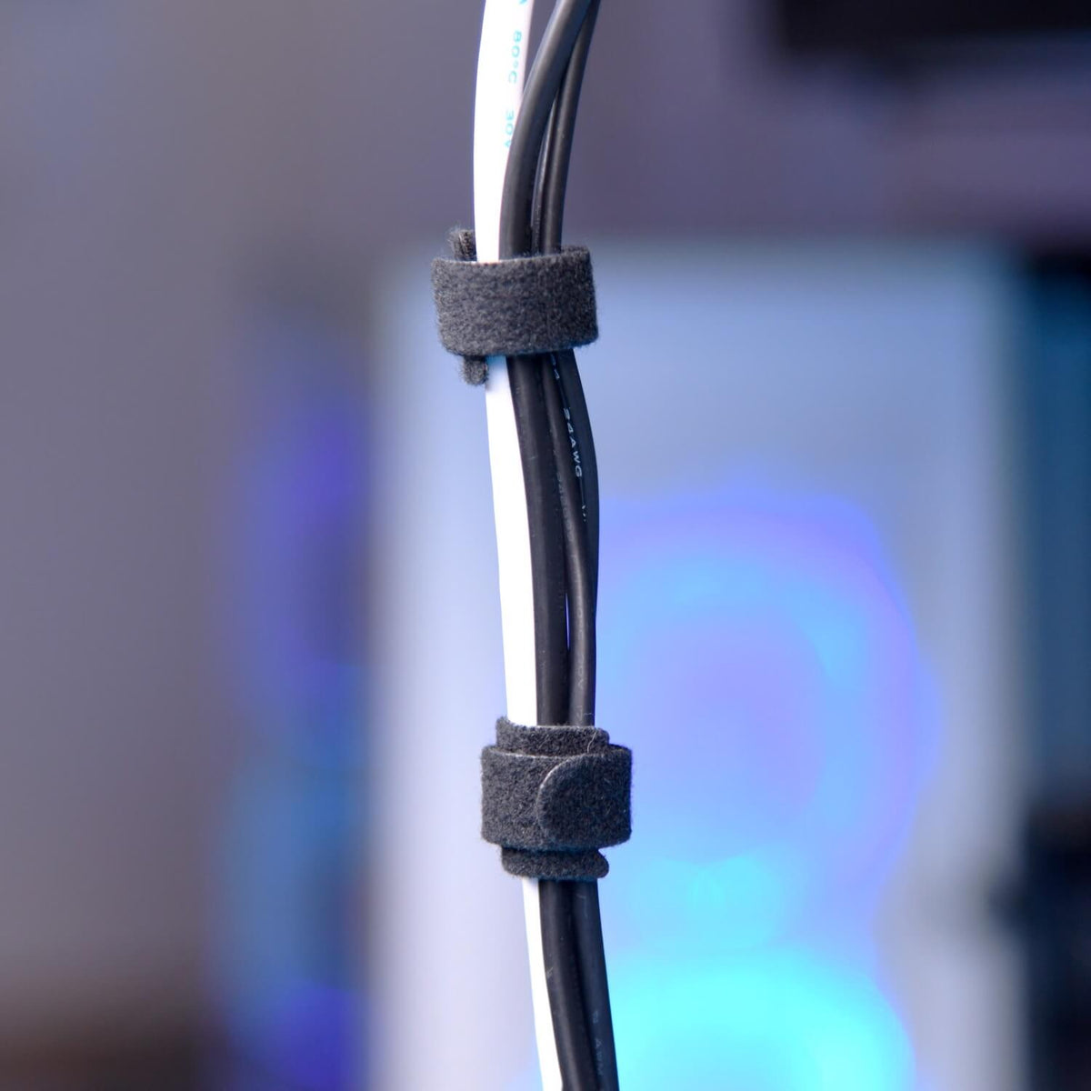 Three black Velcro cable ties secure white and black cables together vertically, with a blurred background showing blue and purple tones, likely from a computer setup.